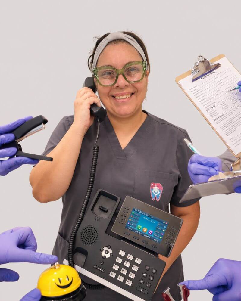 A smiling patient coordinator at Love Your Jaws Surgery Center in South Miami wearing grey scrubs and glasses, holding an office phone and surrounded by gloved hands holding administrative supplies.