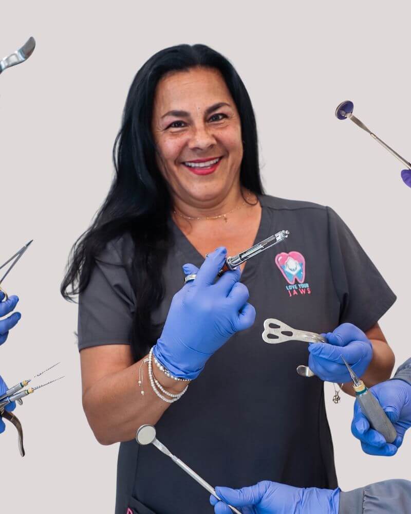 A smiling surgical assistant at Love Your Jaws Surgery Center in South Miami wearing grey scrubs and blue gloves, holding a dental syringe among hands displaying various oral surgery instruments.