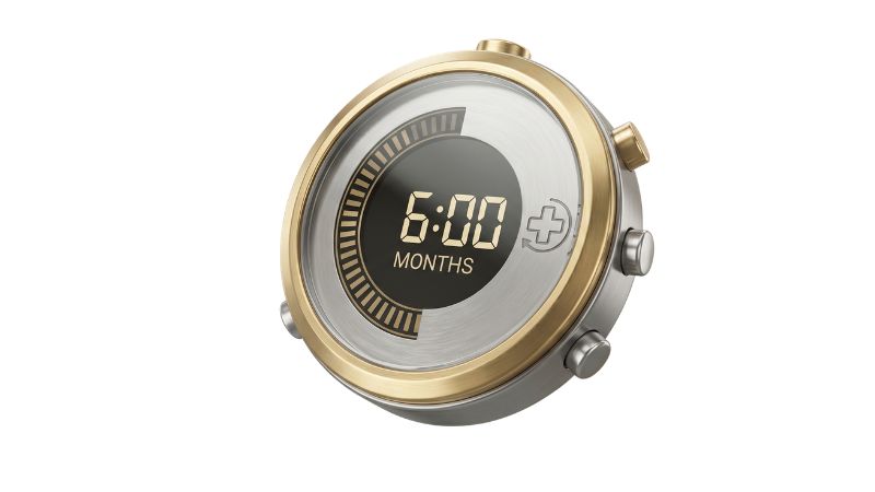 A sophisticated, single 3D medical-grade treatment timer made of brushed titanium with a polished gold alloy bezel. The central illuminated digital display clearly reads "6:00 MONTHS," representing the treatment duration, set against a softly blurred, warm clinical dental office background that perfectly matches the perspective and style of the cost pictogram.