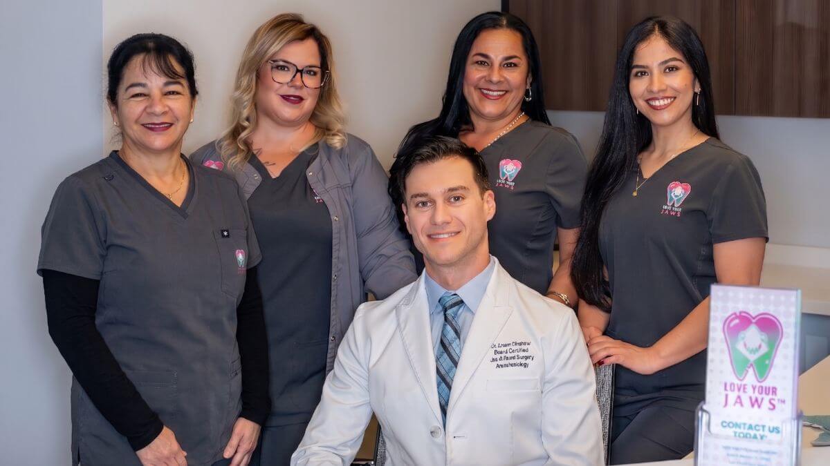 A male oral surgeon in a white lab coat sits at a desk, surrounded by four smiling female dental staff members wearing grey Love Your Jaws scrubs in a professional clinic setting.