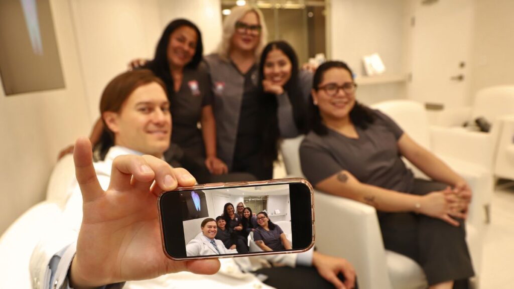 A male oral surgeon in a white lab coat holds up a smartphone to take a group selfie with five female clinic staff members. The camera focuses on the phone screen, which clearly displays the smiling team inside a brightly lit, modern dental office.