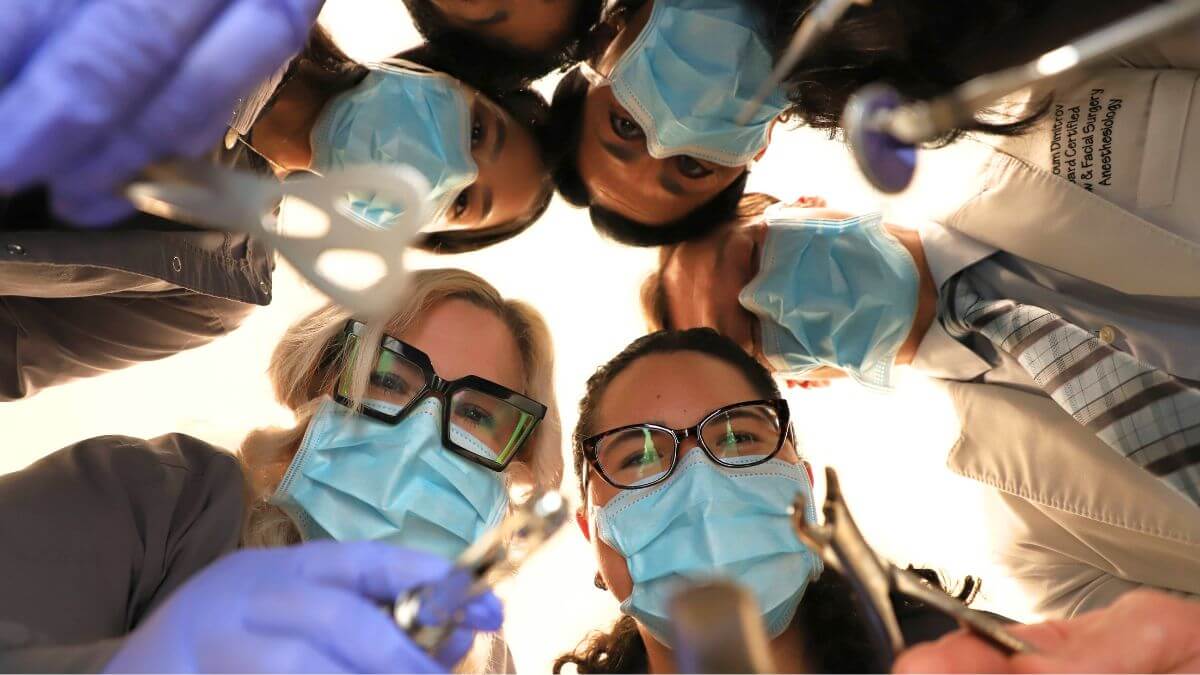 A patient's point-of-view shot looking up at a team of six dental professionals. They are wearing blue surgical masks and holding dental tools, peering down as if leaning over a dental chair.