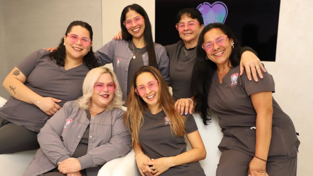 Group portrait of six smiling female dental staff members. They are wearing matching grey scrubs featuring the Love Your Jaws logo and playful translucent pink heart-shaped sunglasses, posing together on white chairs in a modern office environment.